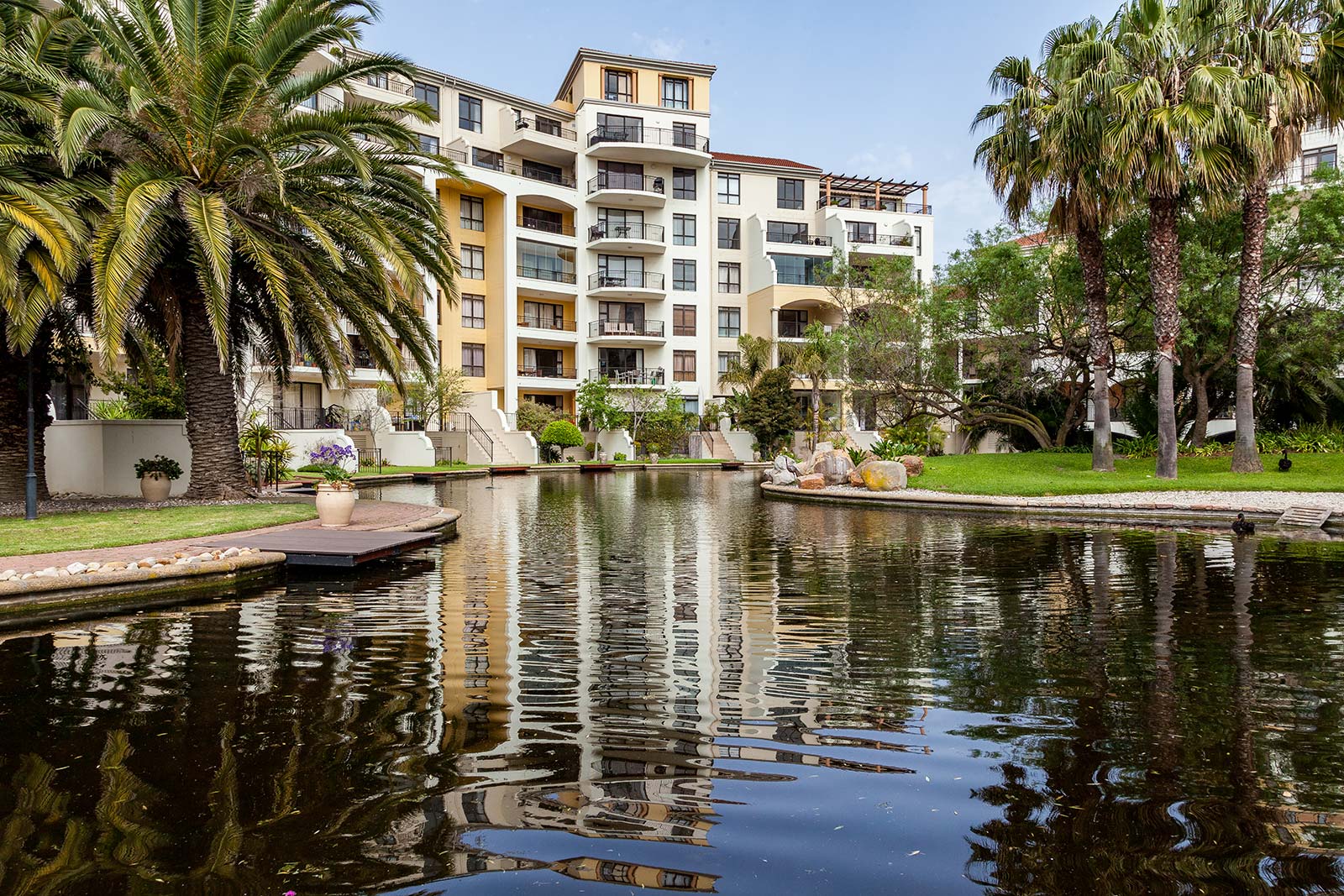 A modern multi-story apartment building by Singer Group features balconies overlooking a landscaped pond surrounded by palm trees and greenery, with clear reflections of the scene shimmering on the water.