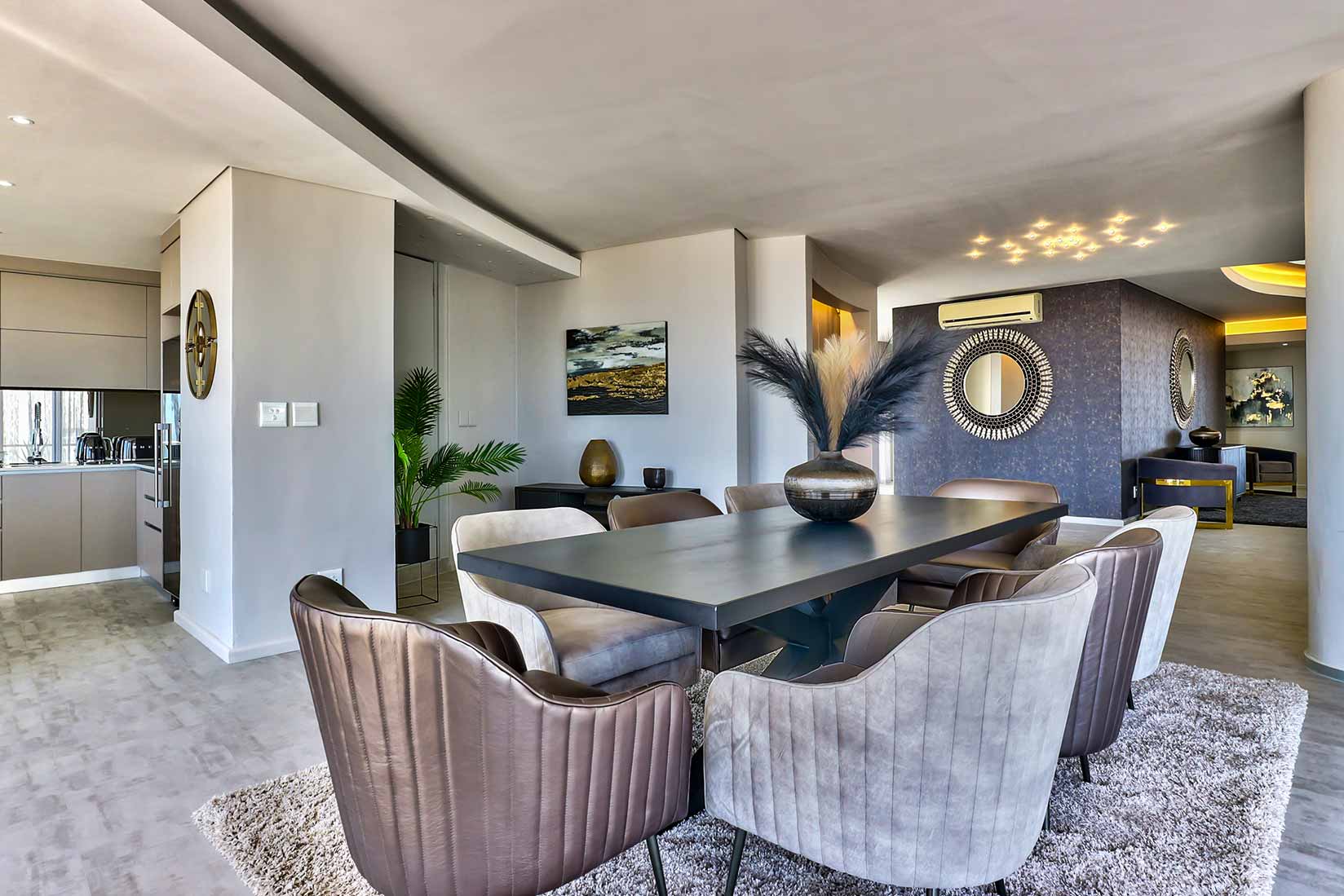 Modern dining room by Singer Group with a sleek black table, six upholstered chairs, a vase with decorative branches, and contemporary art and mirrors on the walls. Neutral tones and abundant natural light fill the space.