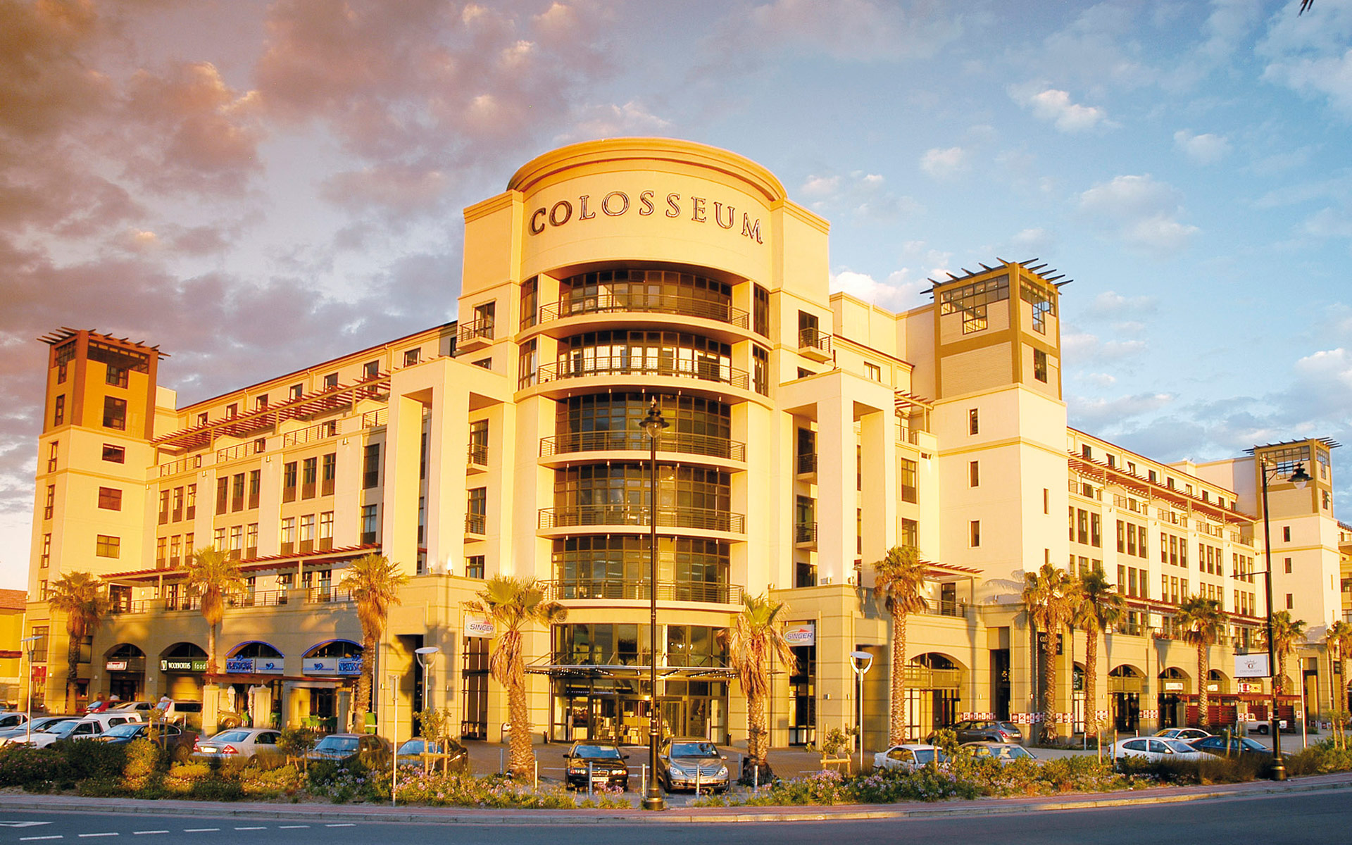A large, modern building with the word "Colosseum" at the top, featuring tall columns, large windows, palm trees in front, and parked cars along the street—home to spectacular Singer Group performances—bathed in golden sunlight.