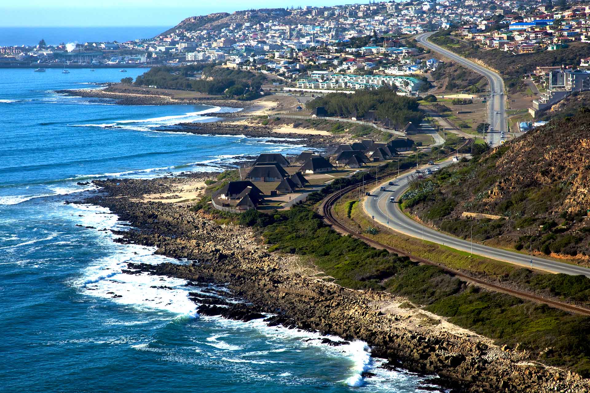 A coastal town with houses and roads running alongside a rocky shoreline; waves crash against the shore near the Singer Group, and a hilly residential area rises in the background under a clear blue sky.