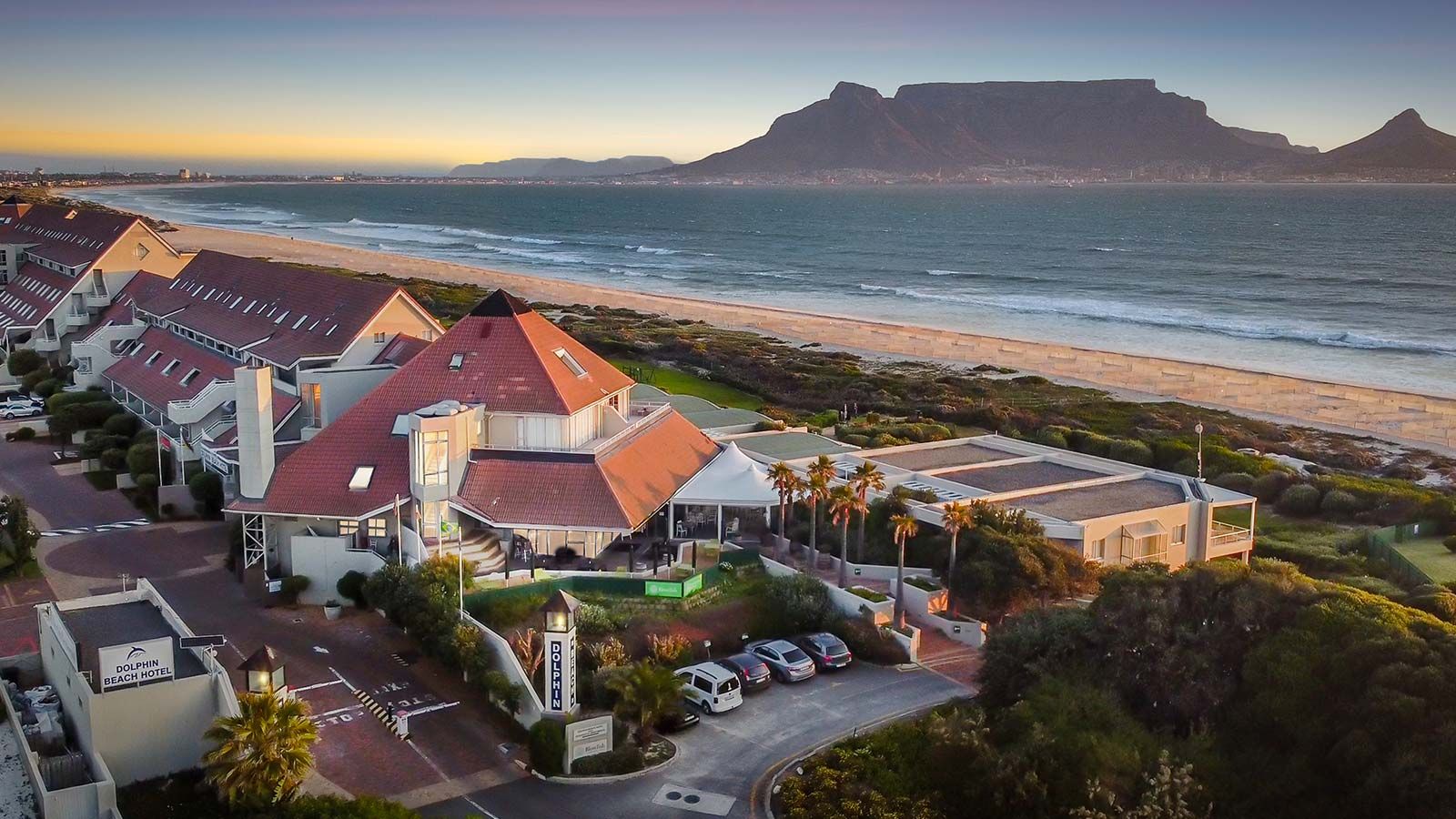 A Singer Group coastal hotel with red-roofed buildings and palm trees sits beside a sandy beach, with rolling waves and Table Mountain visible in the background under a clear sky at sunset.