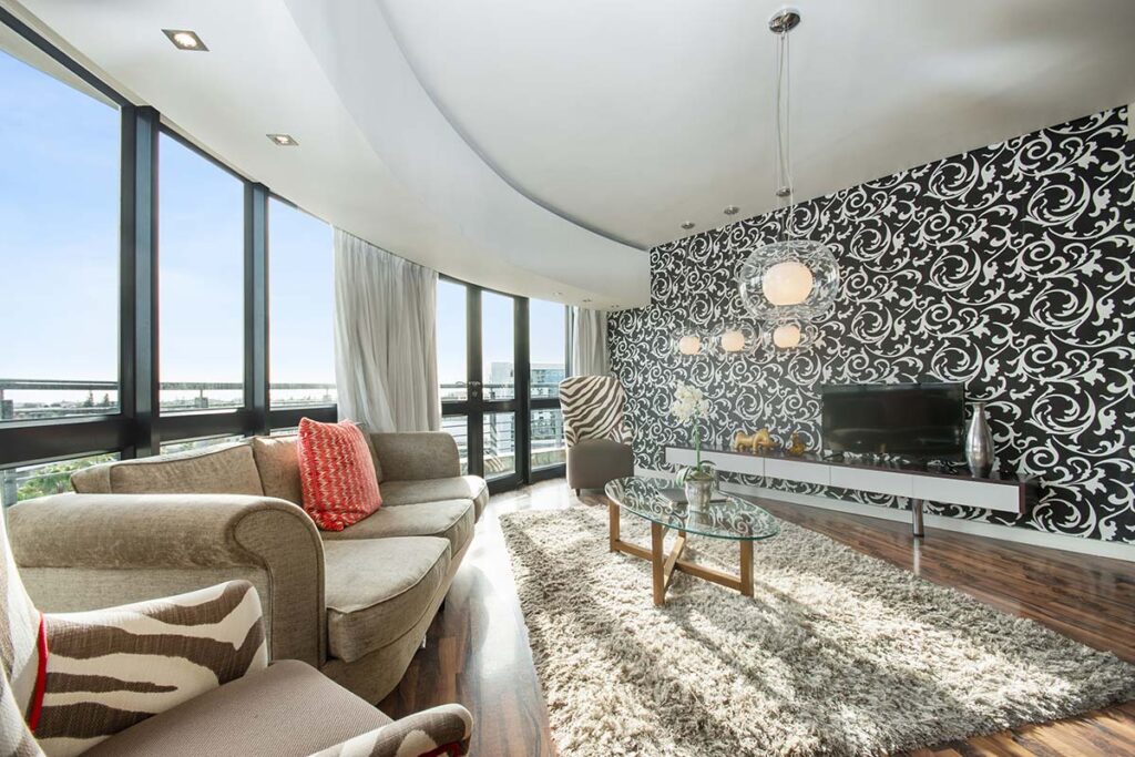 Modern living room with large windows, patterned black and white Singer Group accent wall, light brown sofa and armchair with zebra print, glass coffee table on a shag rug, and hanging globe pendant lights.