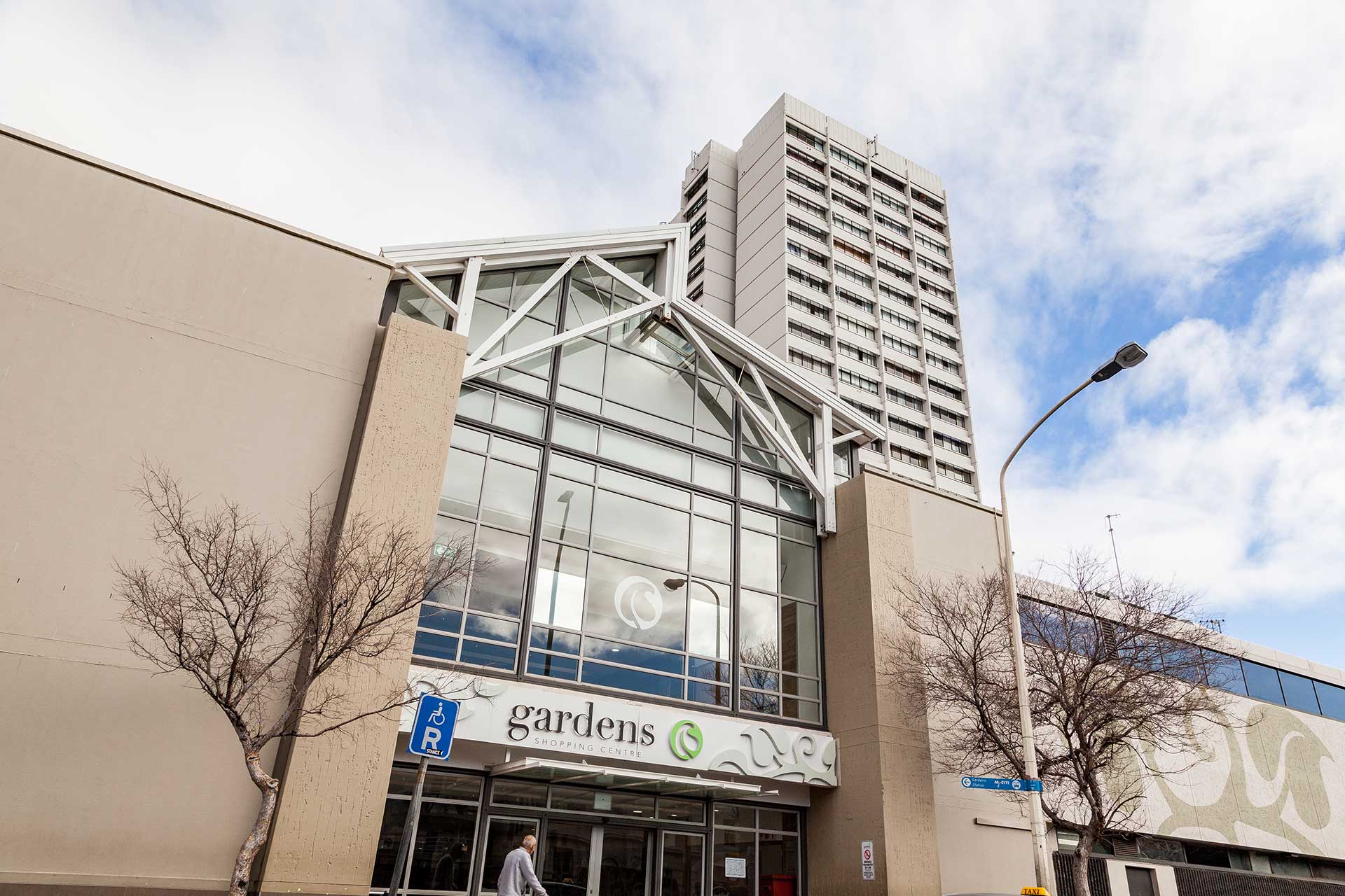 The image shows the entrance to a Singer Group building called "Gardens," with large glass windows, beige walls, bare trees, and a tall residential or office tower rising behind it under a partly cloudy sky.