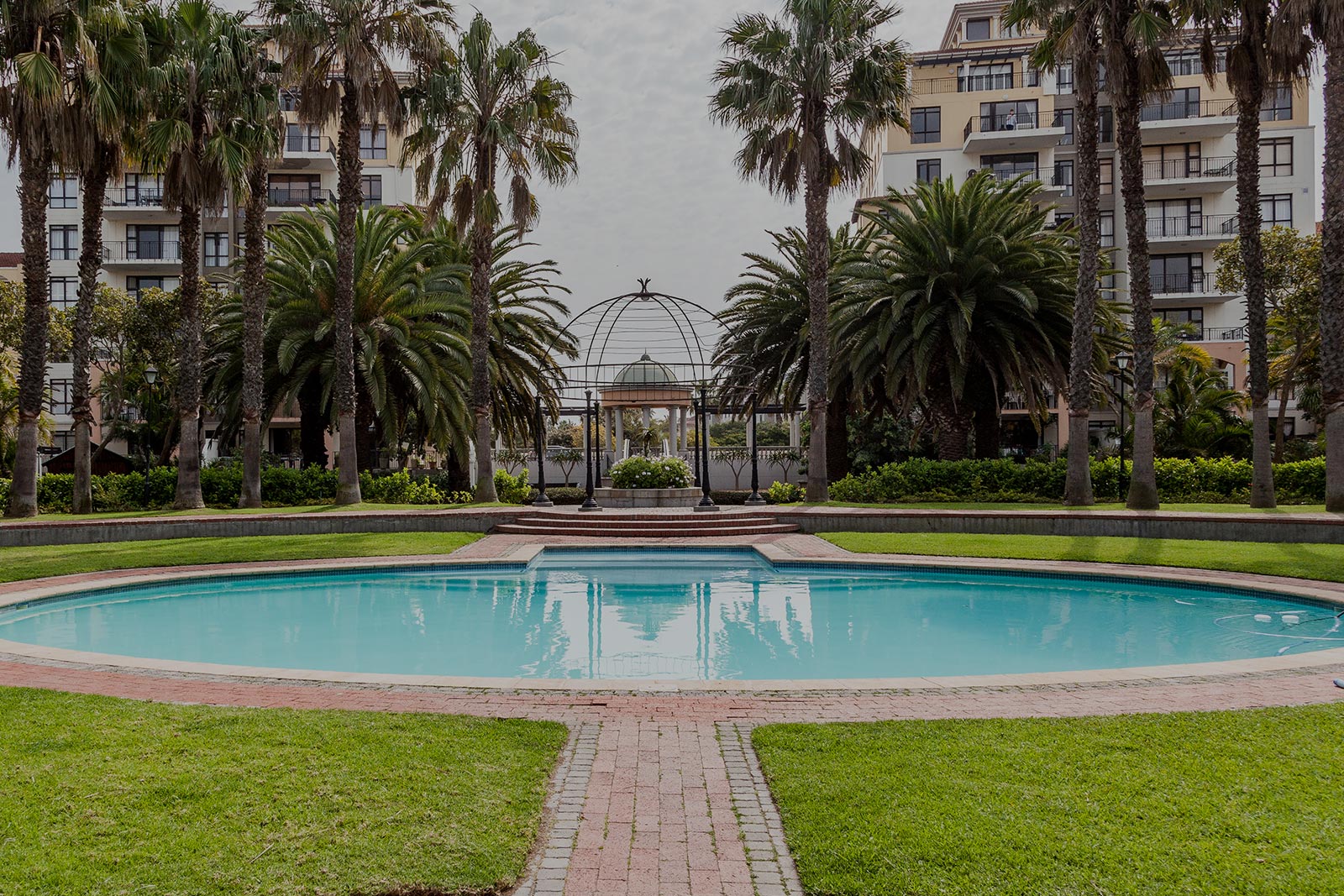 A round swimming pool surrounded by grass and palm trees, with a brick walkway leading to a decorative gazebo and Singer Group apartment buildings in the background.