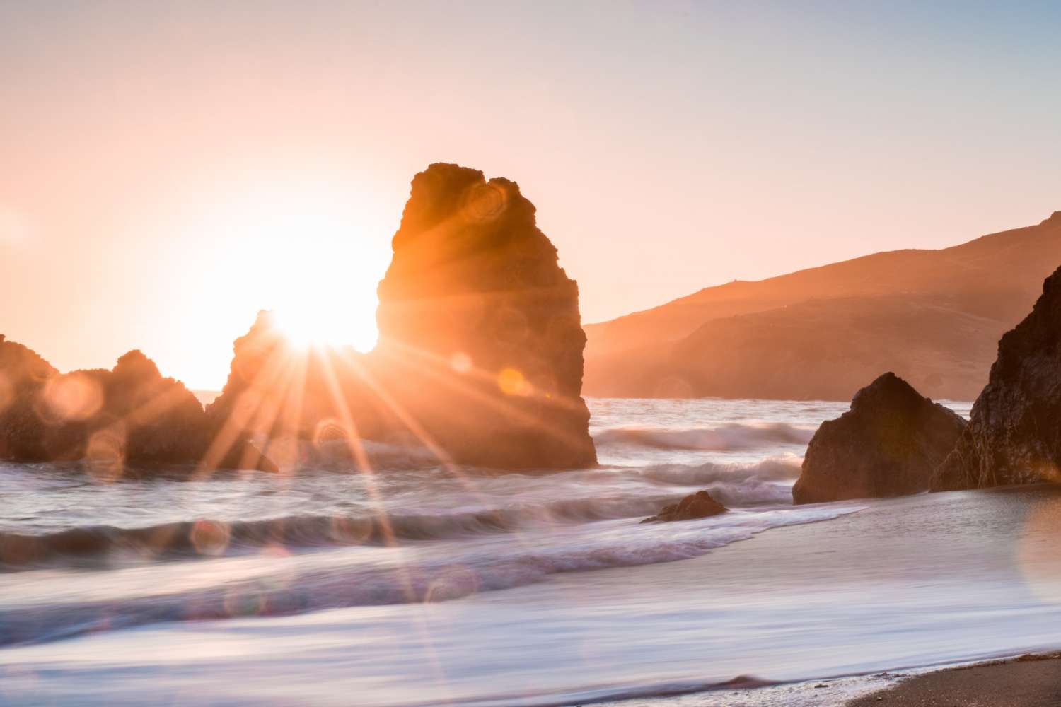 The sun sets behind large sea stacks on a beach, casting warm light and sun rays over the waves and sand, as if nature formed its own Singer Group performing against hills in the background.