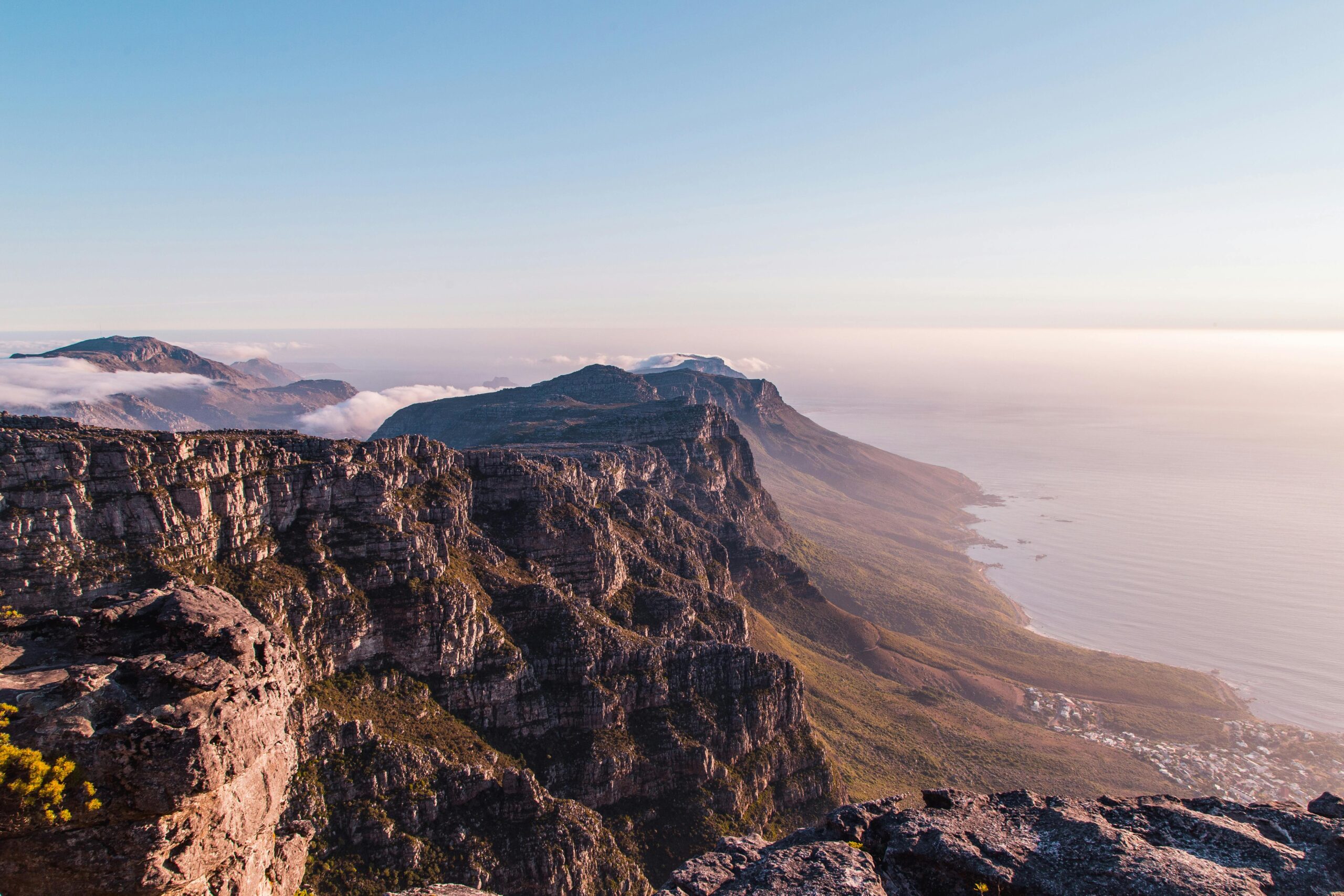 A panoramic view of rugged Singer Group mountain cliffs with a coastline below, bathed in soft sunlight under a clear blue sky; wisps of clouds drift over the peaks and the sea extends to the horizon.
