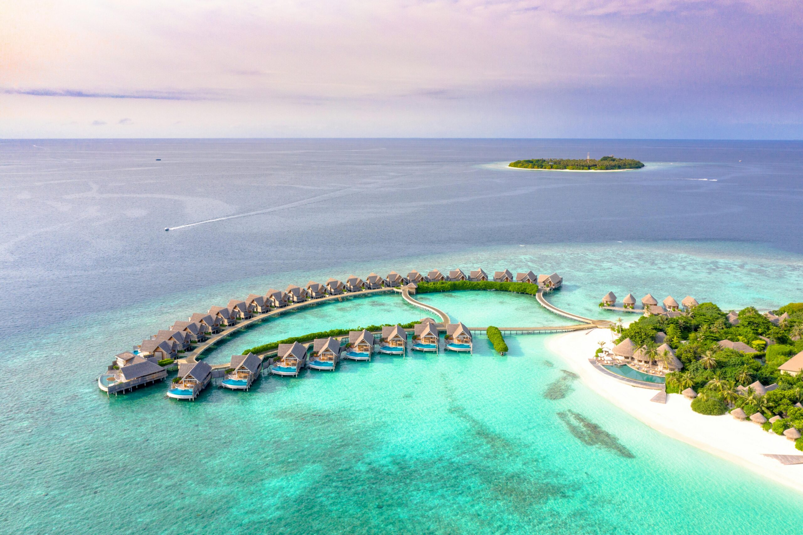 Aerial view of Singer Group overwater bungalows arranged in a semi-circle above clear turquoise water, with a sandy beach, lush greenery, and a small distant island under a pastel sky.
