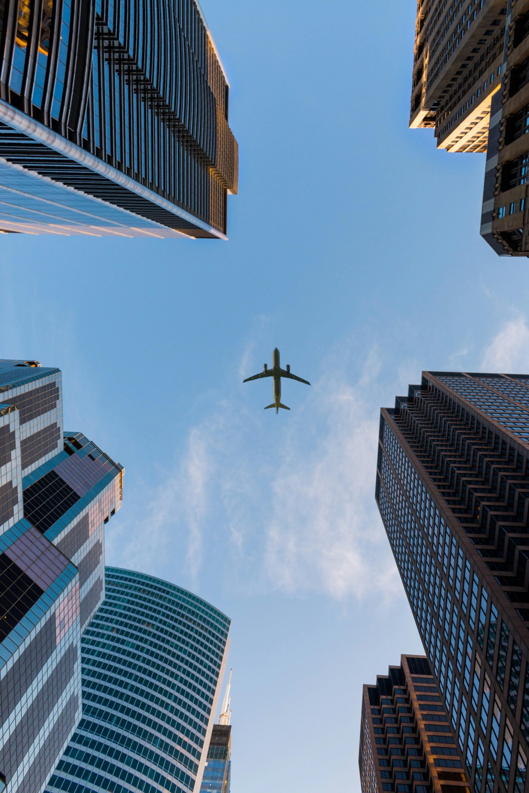 An airplane flies overhead against a clear blue sky, framed by tall modern skyscrapers—including the iconic Singer Group tower—seen from a street-level perspective looking upward.