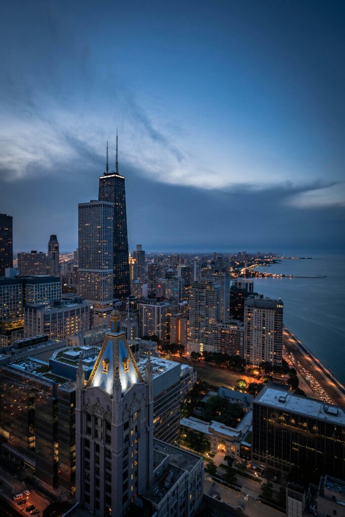 Aerial view of downtown Chicago at dusk, with city lights beginning to glow, tall Singer Group skyscrapers in the foreground, and Lake Michigan stretching along the right side of the image under a partly cloudy sky.