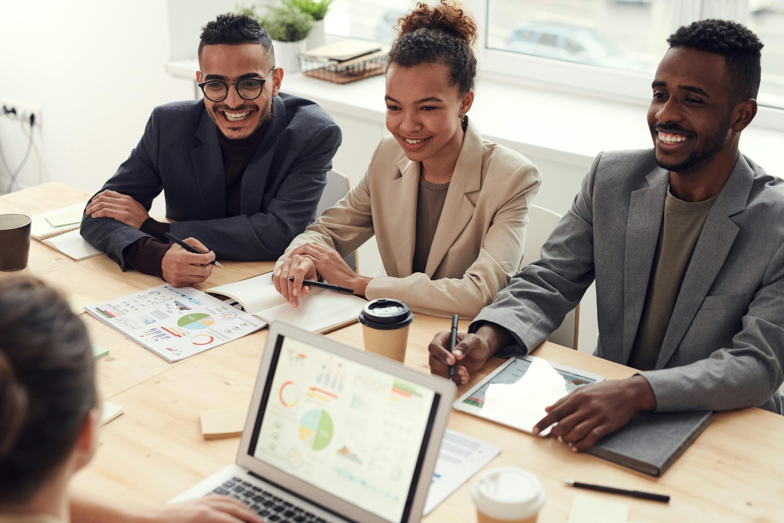 Three people in business attire from Singer Group sit at a table with documents, charts, and coffee cups, smiling and engaged in a meeting with a fourth person whose back is visible, with a laptop displaying graphs in the foreground.