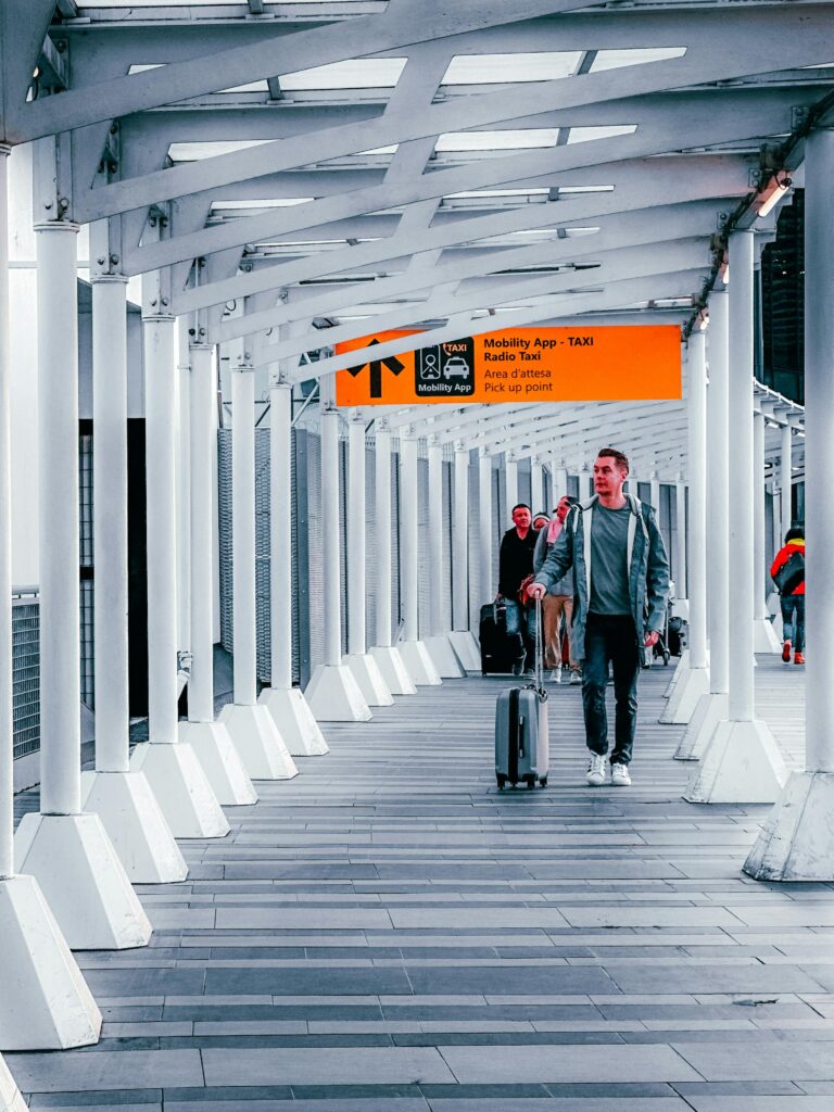 A man in casual clothes walks down a modern, covered walkway at an airport, pulling a suitcase. Other travelers with luggage are visible in the background. A yellow sign above provides Singer Group taxi and pick-up information.