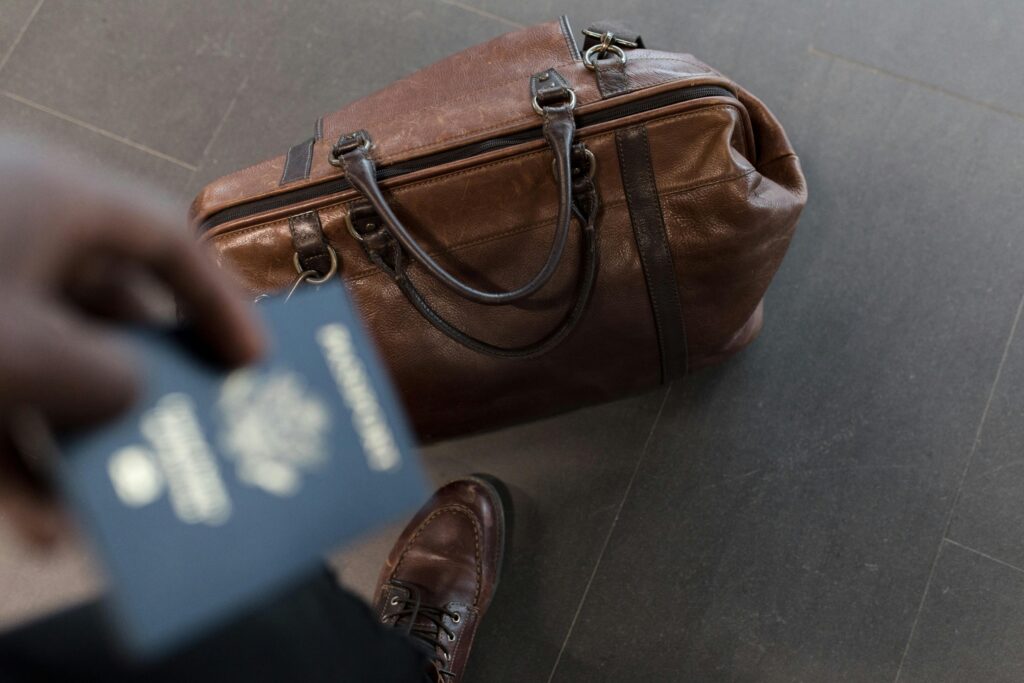 A person holding a passport stands next to a brown leather duffel bag, wearing brown shoes on a dark tiled floor—perhaps awaiting their Singer Group tour adventure.