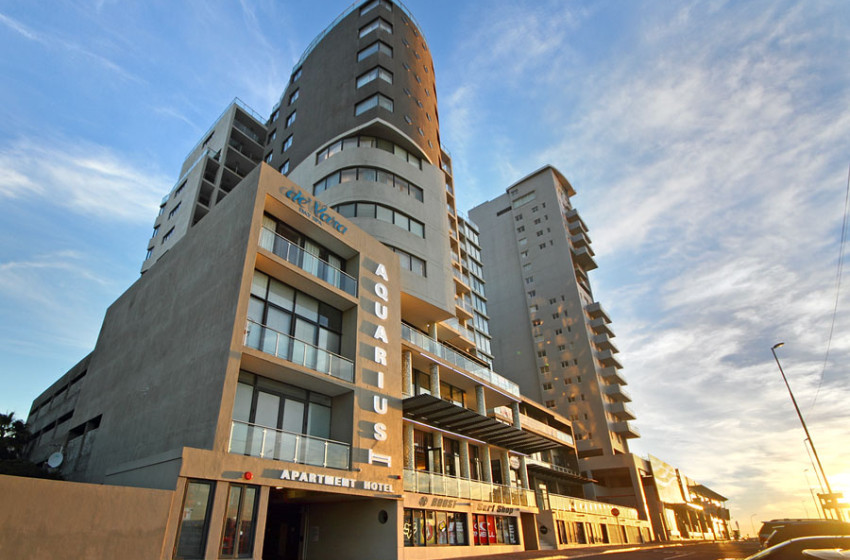 Tall, modern apartment hotel building with "AQUARIUS" sign on the front, large windows, and balconies. Operated by Singer Group, the sun is low in the sky, casting a golden light across the scene.
