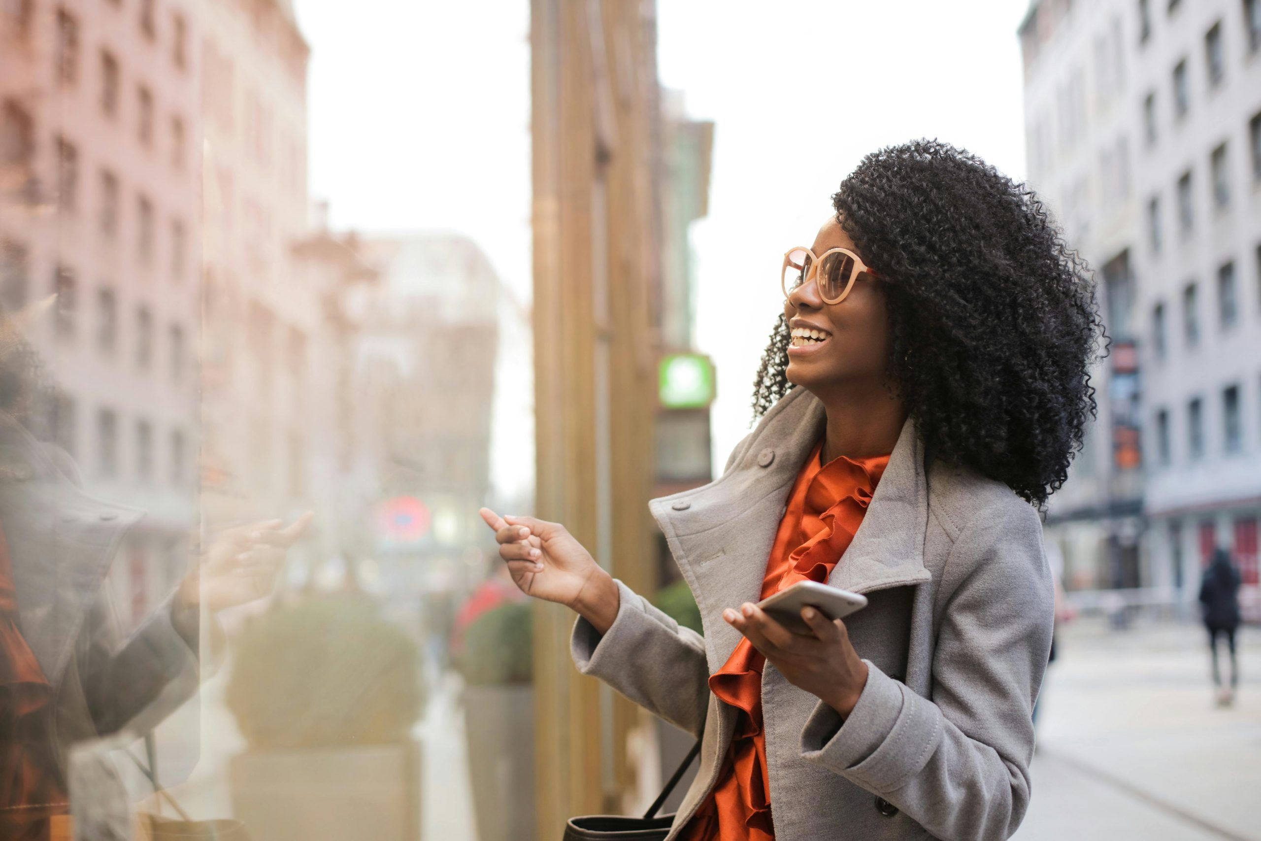 A woman with curly hair, wearing sunglasses and a gray coat, smiles while looking at a shop window and holding her phone on a city street, perhaps heading to meet friends from her singer group.